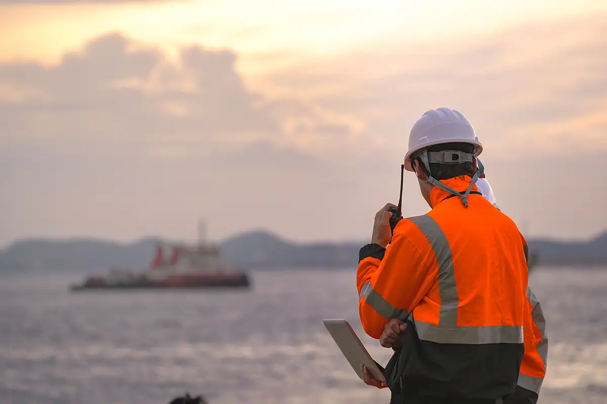 Tarchon Interconnector – Offshore-Betrieb und Projektdokumentation Offshore-Energiearbeiter in einer gut sichtbaren orangefarbenen Jacke und einem weißen Schutzhelm, der ein Tablet oder Funkgerät benutzt; im Hintergrund sind in der Abenddämmerung Schiffe zu sehen.